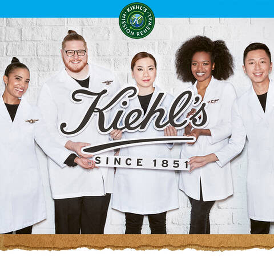 Cinco empleados de Kiehl's sonrientes con batas blancas sosteniendo el logo. Emblema 'Mission Renew