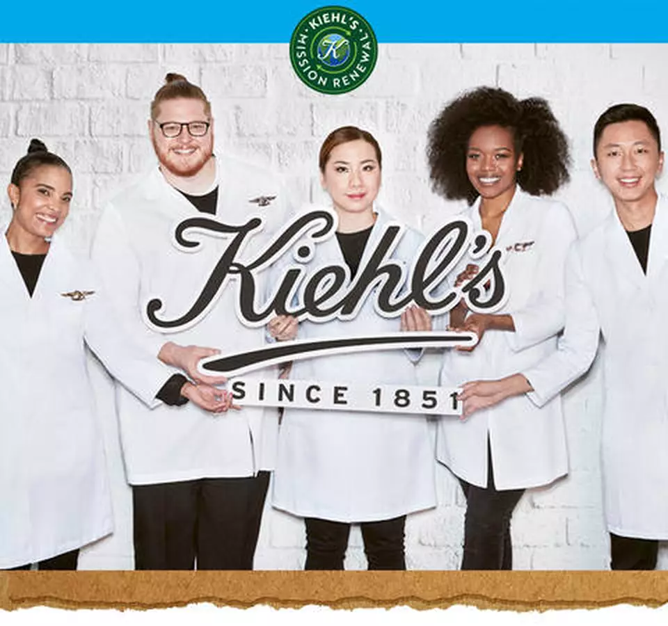 Cinco empleados de Kiehl's sonrientes con batas blancas sosteniendo el logo. Emblema 'Mission Renew